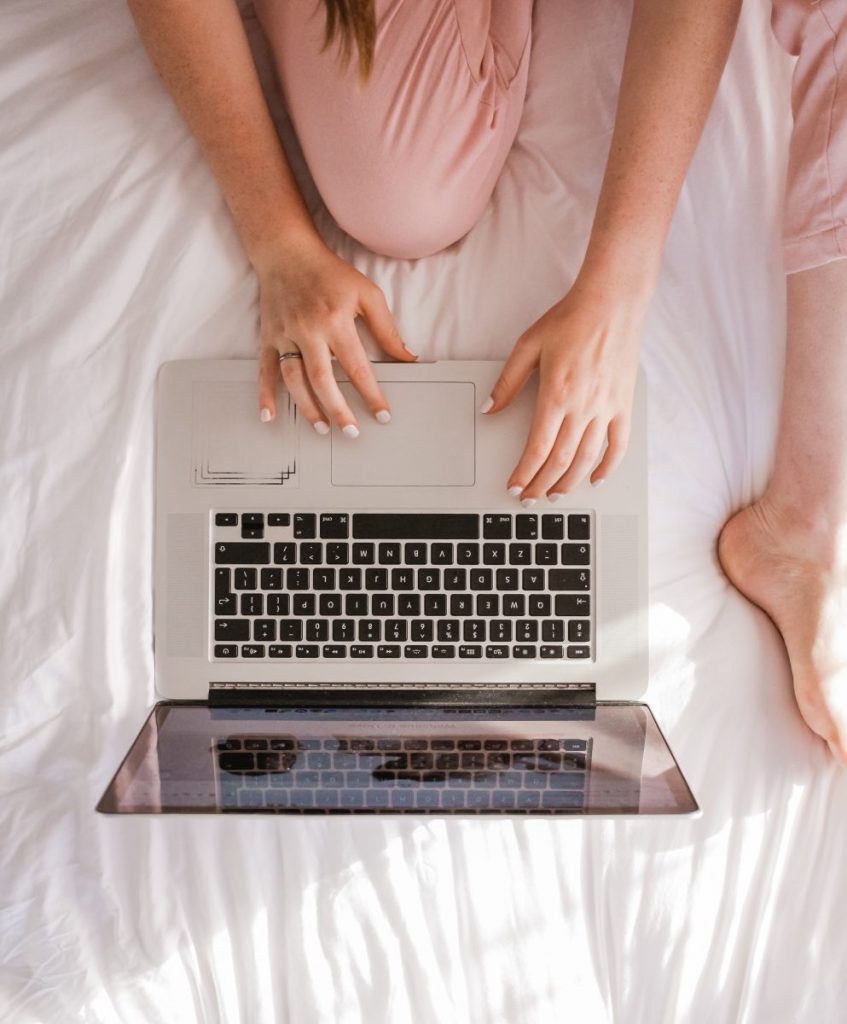 Photo of a woman comfortably sitting on a bed with a laptop.